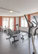 Lichtdurchfluteter Fitnessraum im Hotel Alpenblick mit modernen Ausdauer- und Kraftgeräten, grossen Fenstern und bereitliegenden gelben Handtüchern.